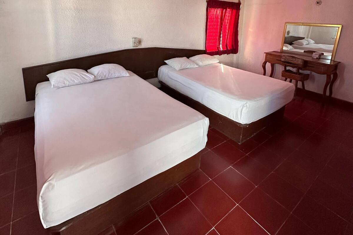 Une chambre d’hôtel avec deux lits simples, un bureau en bois, un ventilateur de plafond et un rideau rouge.