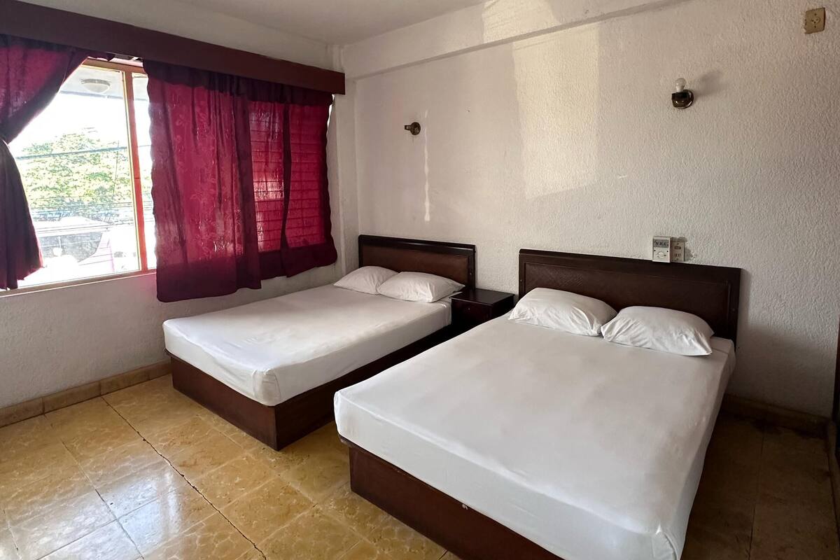 Une chambre avec deux lits simples, un ventilateur de plafond, une fenêtre avec des rideaux rouges et un sol carrelé.