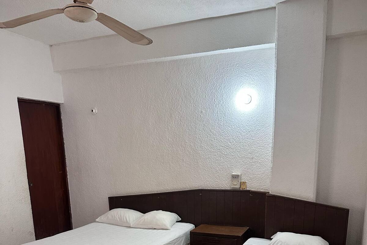 Une chambre avec deux lits simples, une tête de lit en bois, un ventilateur de plafond et un luminaire fixé au mur.