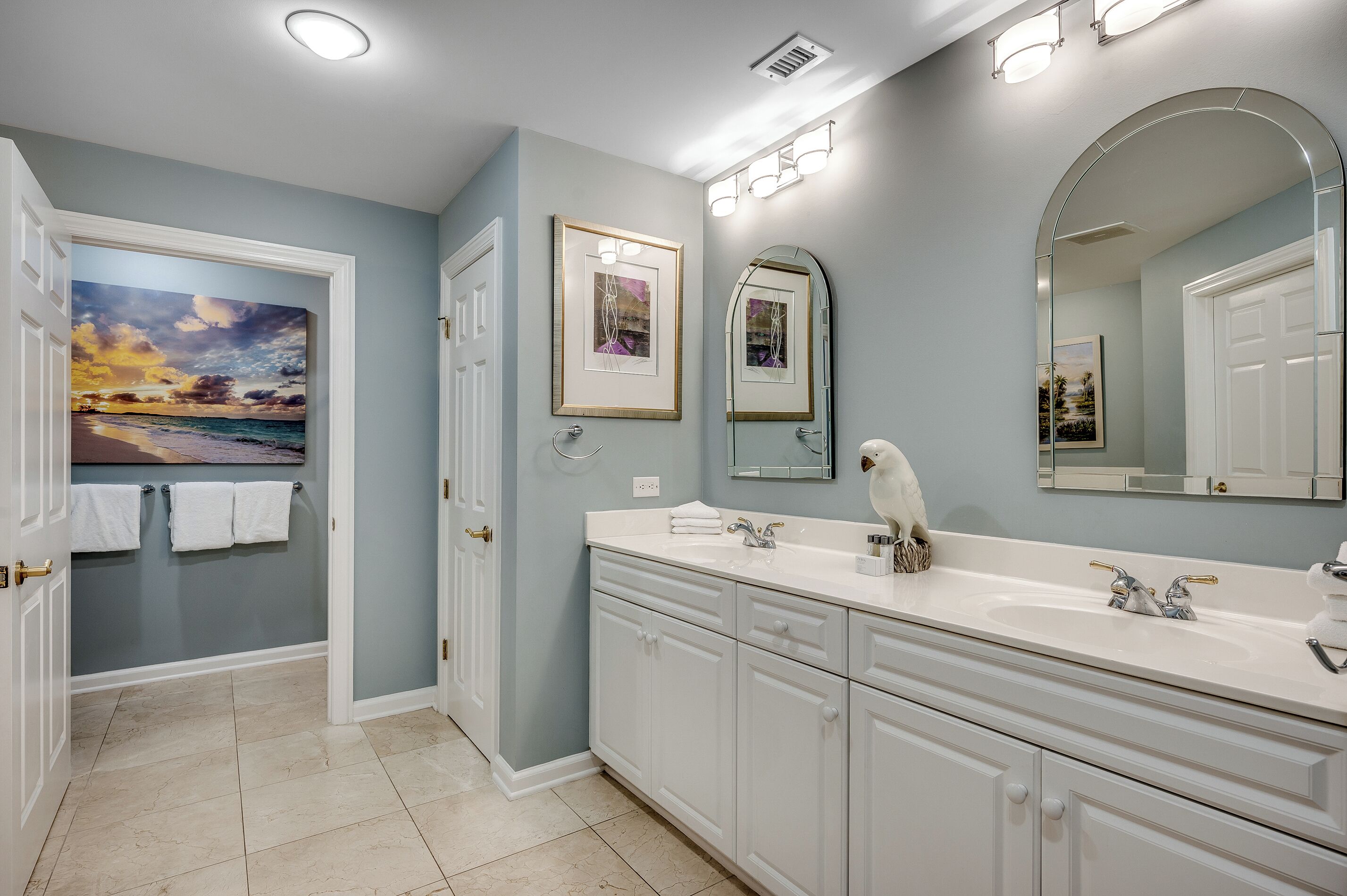 A bathroom with a large mirror, two sinks, and a painting of a sunset on the wall.