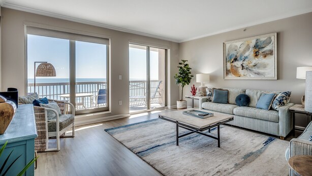 Ein geräumiges Wohnzimmer mit einem großen Schiebeglastür, die zu einem Balkon mit Meerblick führt. Es gibt ein Sofa, einen Couchtisch und einen Beistelltisch mit einer Lampe.