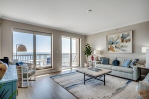 A spacious living room with a large sliding glass door leading to a balcony with a view of the ocean. There is a sofa, a coffee table, and a side table with a lamp.