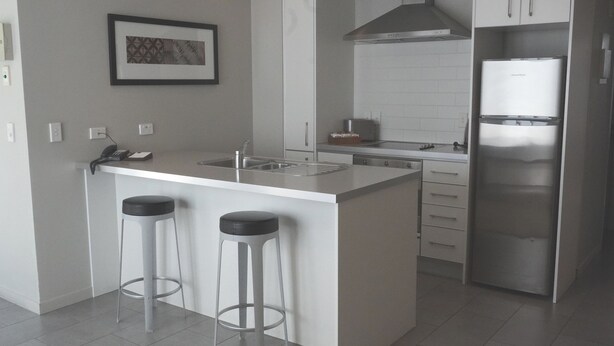 A modern kitchen with a central island, two bar stools, a stainless steel refrigerator, and a tiled backsplash. - Paihia Waterfront 1 Bedroom Apt & Pool (Paihia)