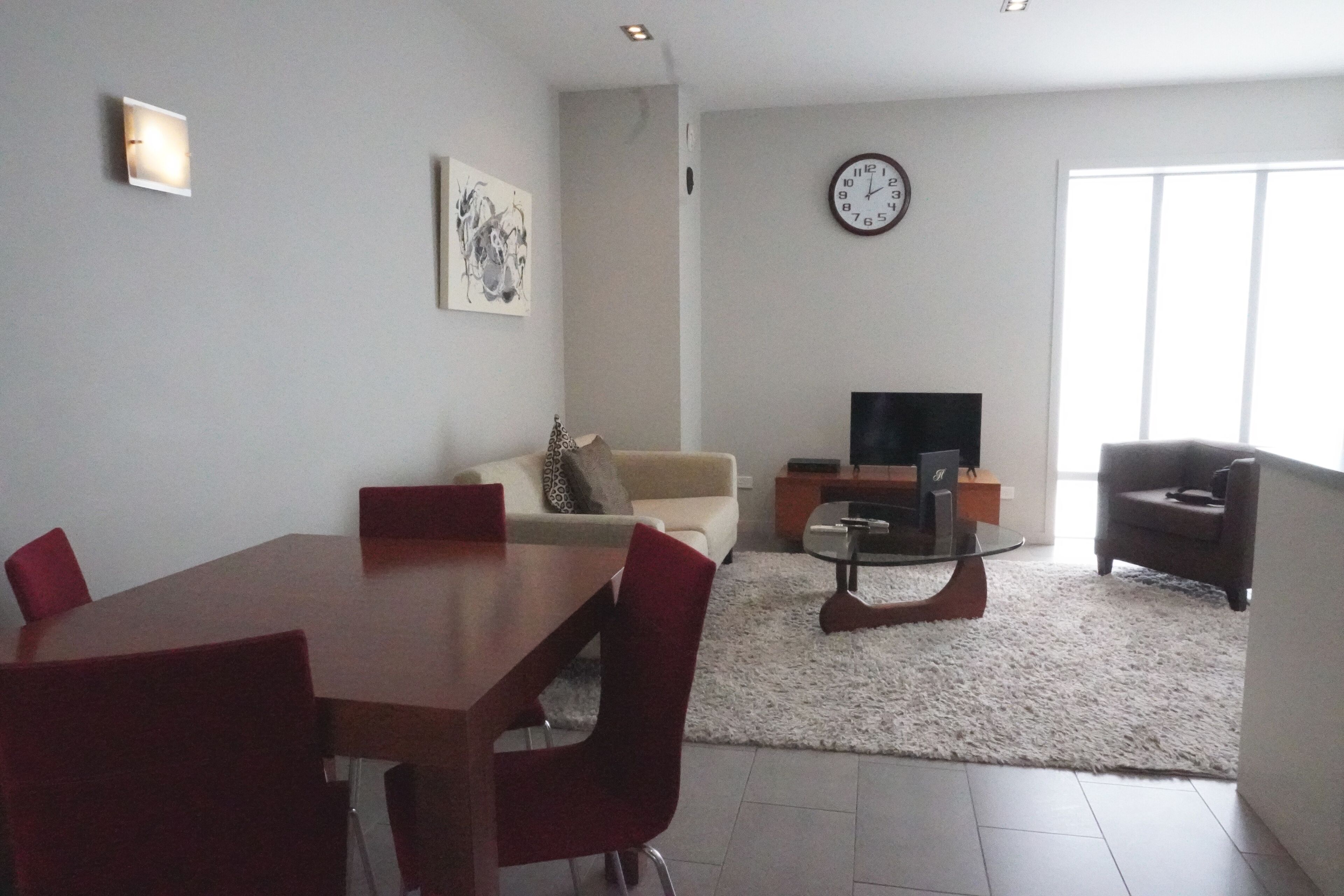 Una sala de estar moderna con una mesa de comedor, sillas rojas, un sofá, un televisor y un reloj en la pared.
