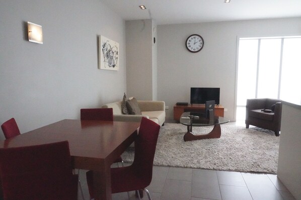 A modern living room with a dining table, red chairs, a sofa, a TV, and a clock on the wall. - Paihia Waterfront 1 Bedroom Apt & Pool (Paihia)