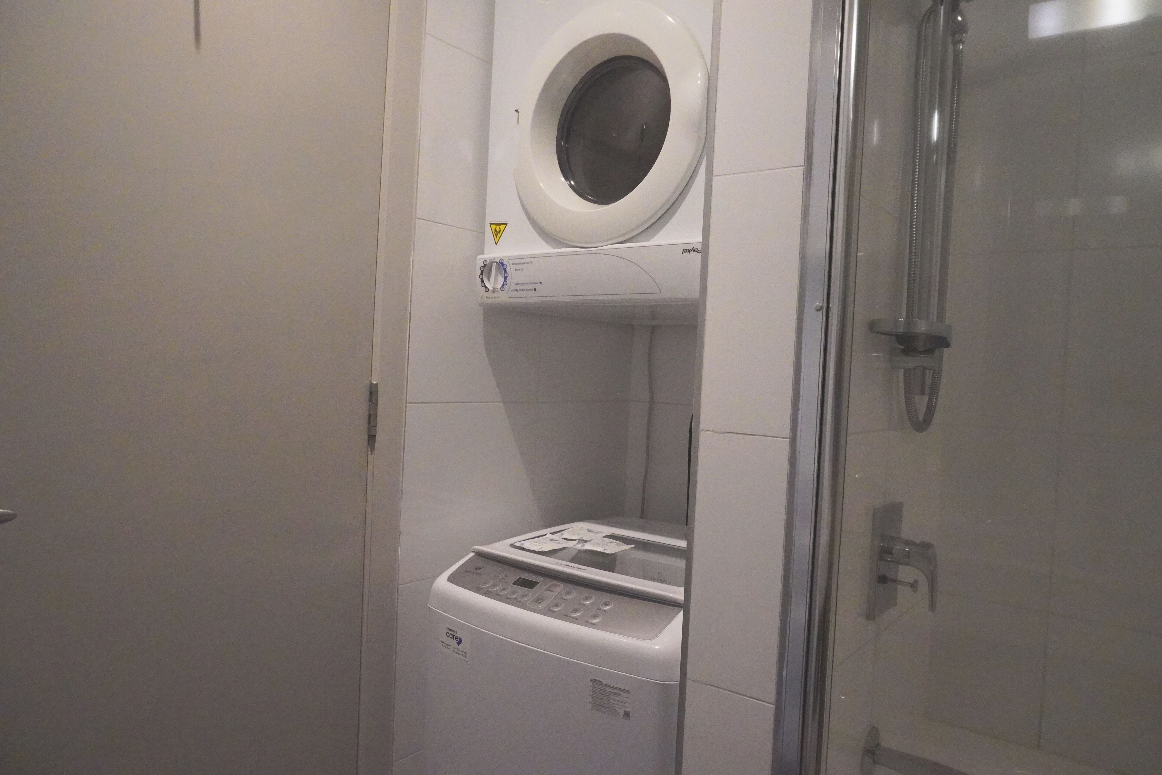Un baño compacto con lavadora y secadora integradas, ducha con mampara de vidrio y una puerta a la izquierda.
