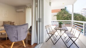 A balcony with a table and chairs, a grey chair, and a sliding glass door leading to an indoor living area with a sofa and a coffee table.