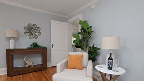 Een woonkamer met een witte fauteuil, een console met een decoratief object, een grote groene plant en een klein rond tafeltje met een wijnfles en glazen.