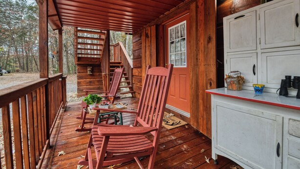 A wooden cabin with a porch, a dining table, chairs, a kitchen area, and a staircase leading to an upper level.