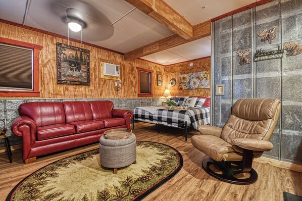 Interior acogedor de una cabaña con un sofá de cuero rojo, un sillón de cuero marrón, un reposapiés redondo y una cama con colcha a cuadros.