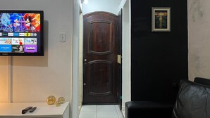 Un salon avec un téléviseur à écran plat diffusant un dessin animé, une porte en bois sombre, un canapé en cuir noir et une petite table sur laquelle sont posées des télécommandes.