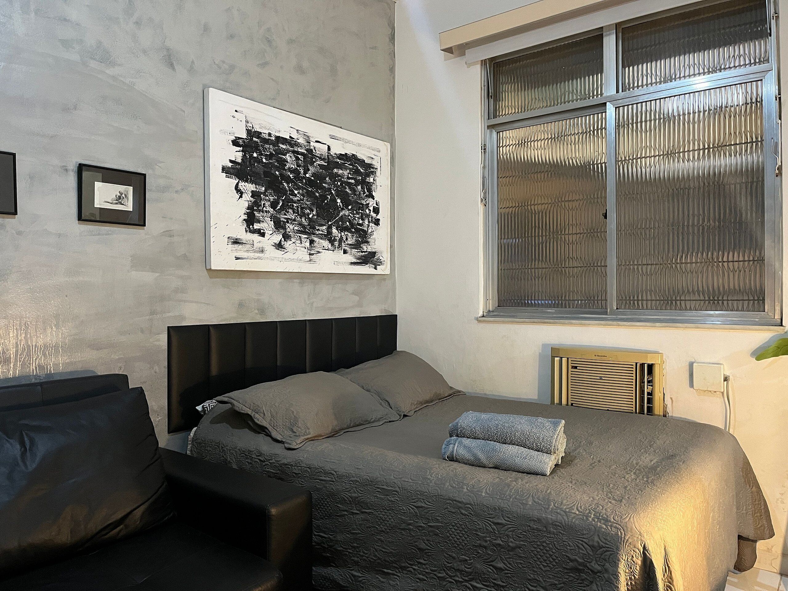 A bedroom with a bed, a headboard, a window with blinds, a radiator, and a framed picture on the wall.