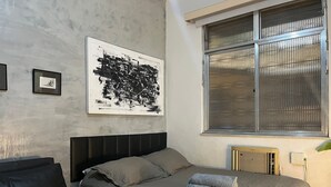 Une chambre à coucher avec un lit, une tête de lit, une fenêtre avec des stores, un radiateur et un tableau encadré au mur.