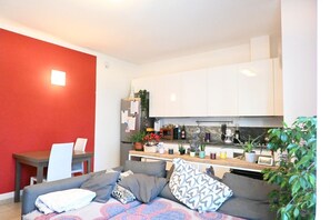 Una sala moderna con un sofá gris, una mesa de comedor con sillas y una cocina con armarios blancos y varios objetos en la encimera.