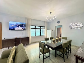 Una sala de estar moderna con televisor de pantalla plana, una mesa de comedor con sillas, un sofá, una araña y un reloj de pared.