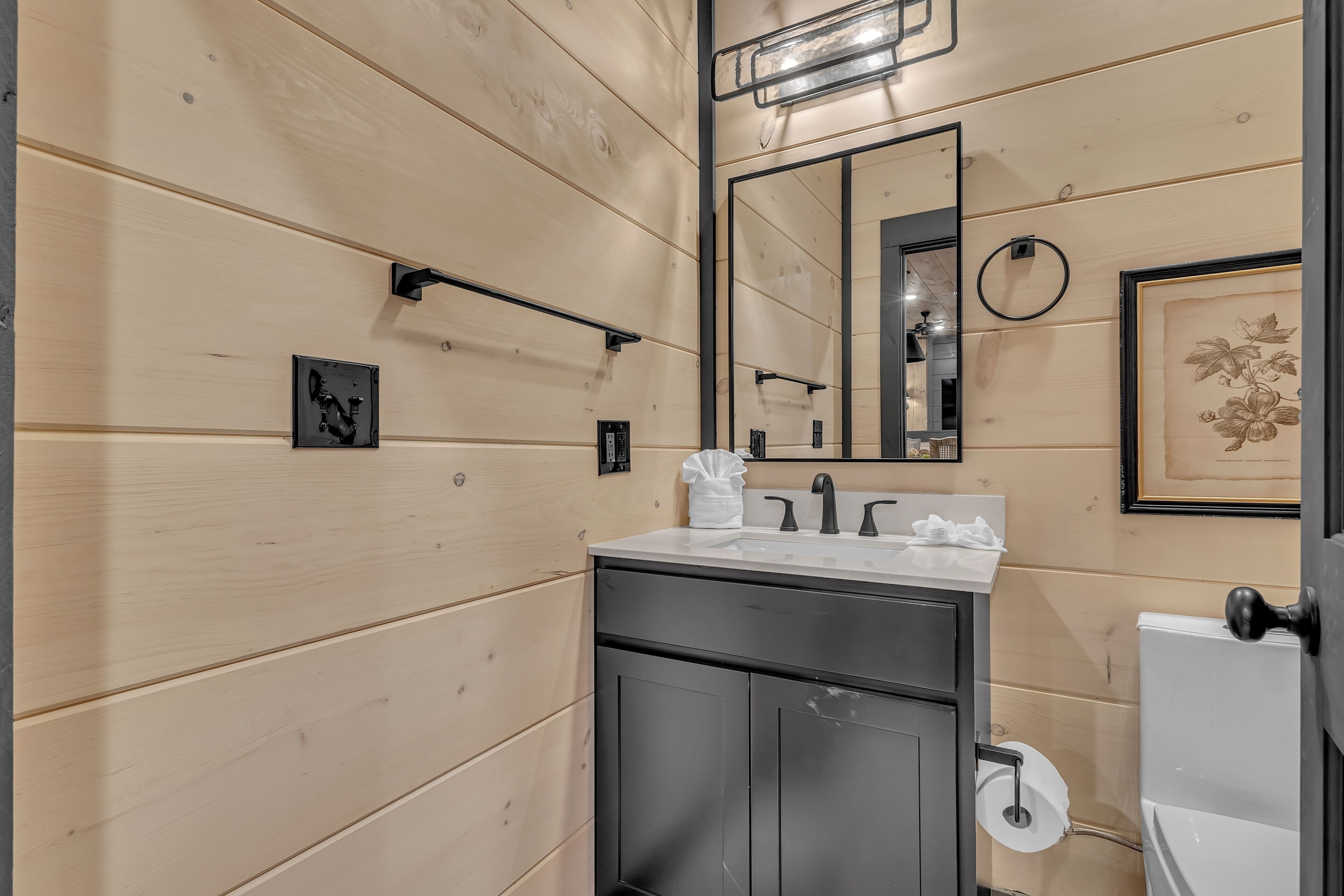 Une salle de bain avec des murs recouverts de panneaux de bois, un lavabo avec un miroir au-dessus, des toilettes et un tableau encadré accroché au mur.