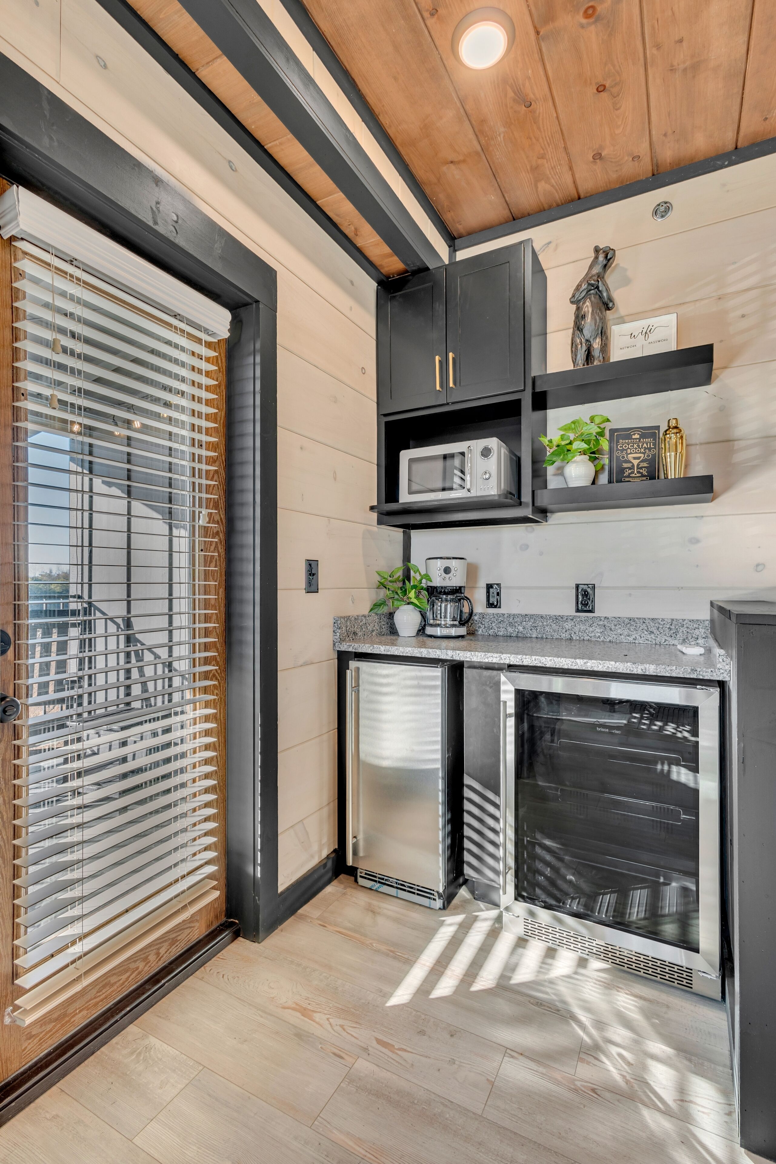 A modern kitchen with built-in appliances, wooden ceiling, and a view through a window with blinds.