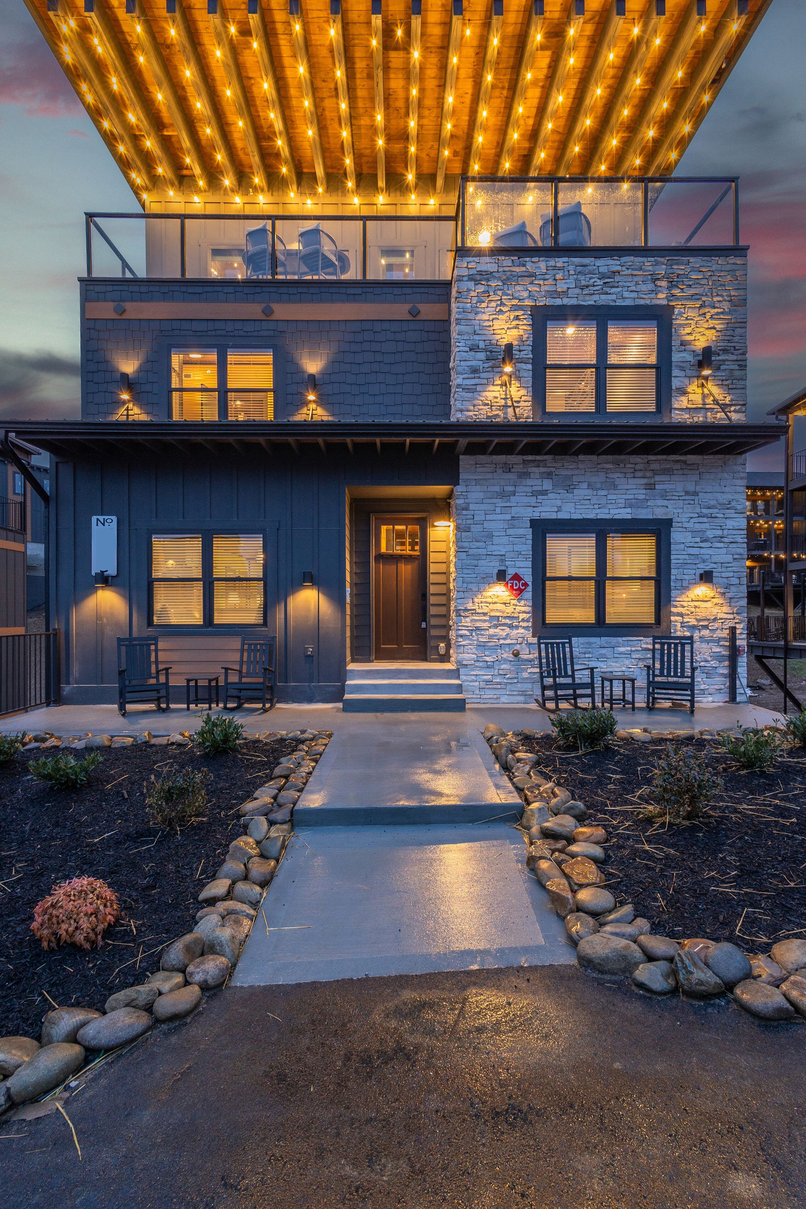 A modern two-story house with a stone facade, a balcony with string lights, and outdoor seating.