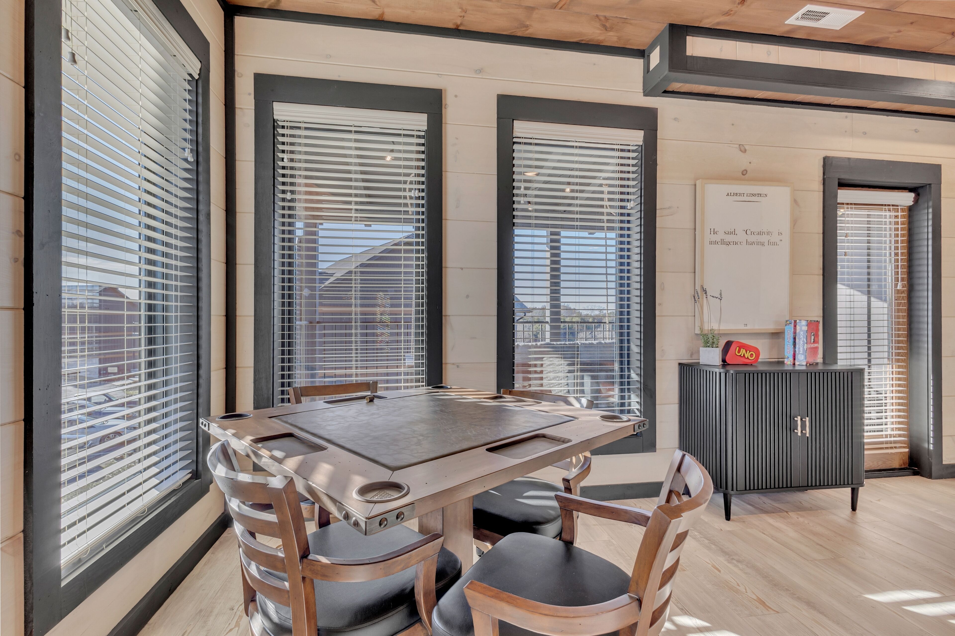 Et moderne spiseområde med et træbord og stole, store vinduer med persienner og et skab med et indrammet citat.
