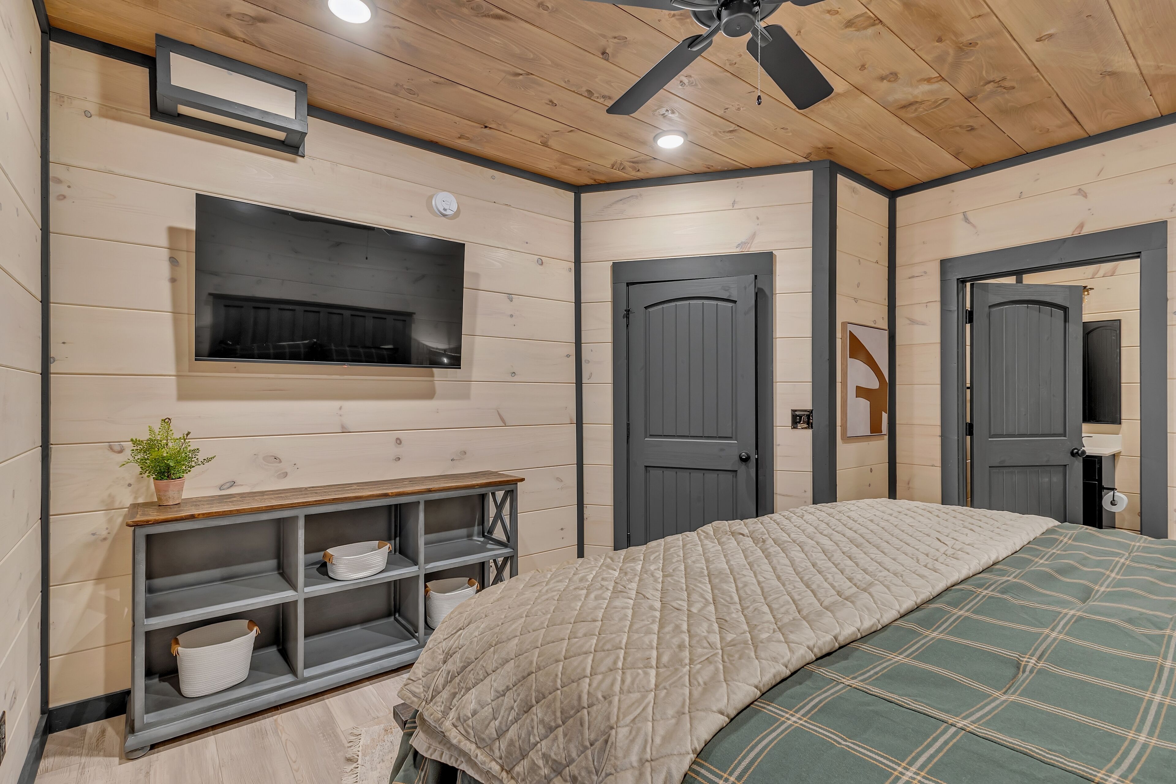 A bedroom with a bed, a TV mounted on the wall, a wooden ceiling, and a door.