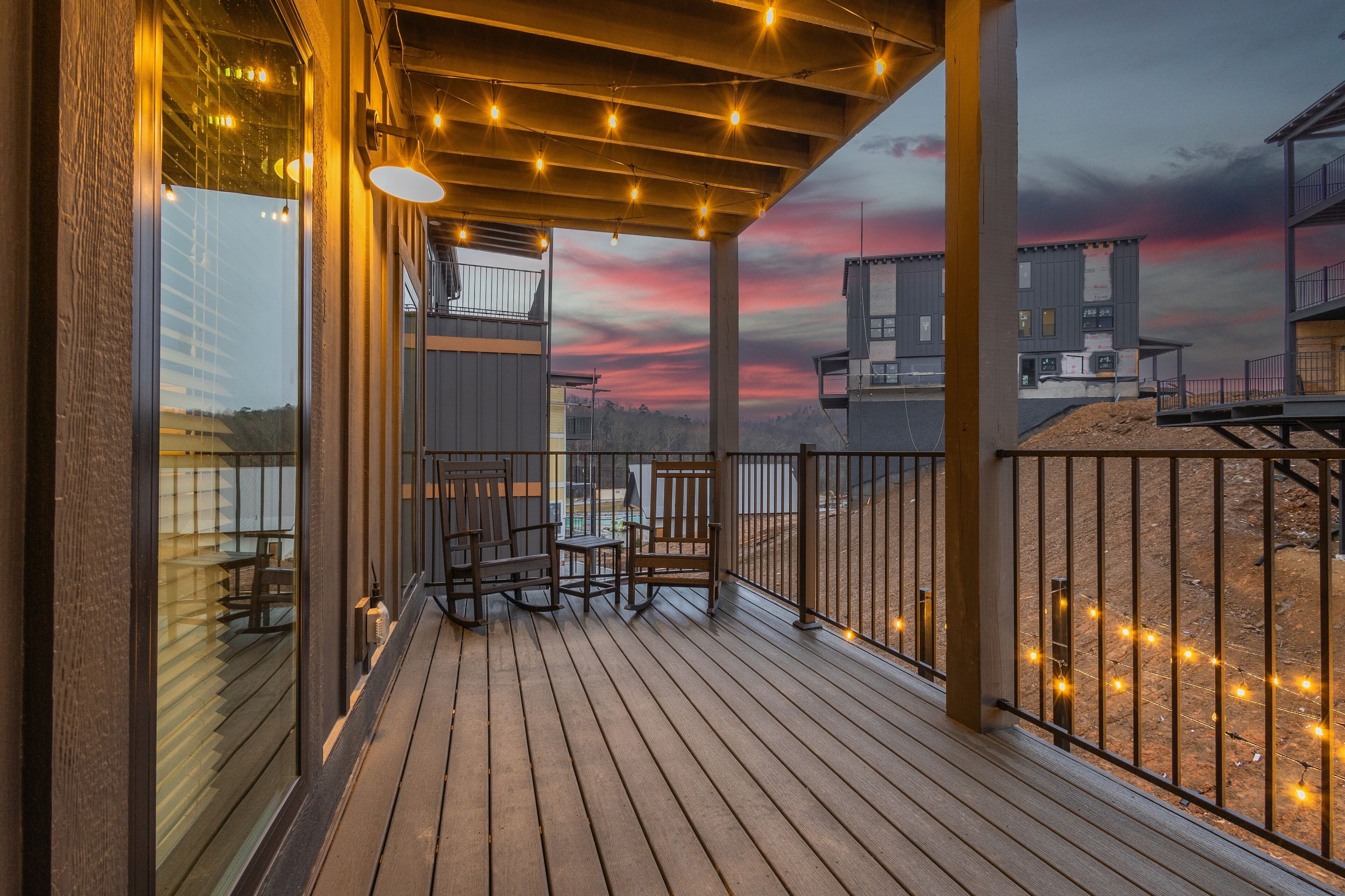 Um deck de madeira com área de estar externa e luzes de corda, com vista para um prédio e um pôr do sol.