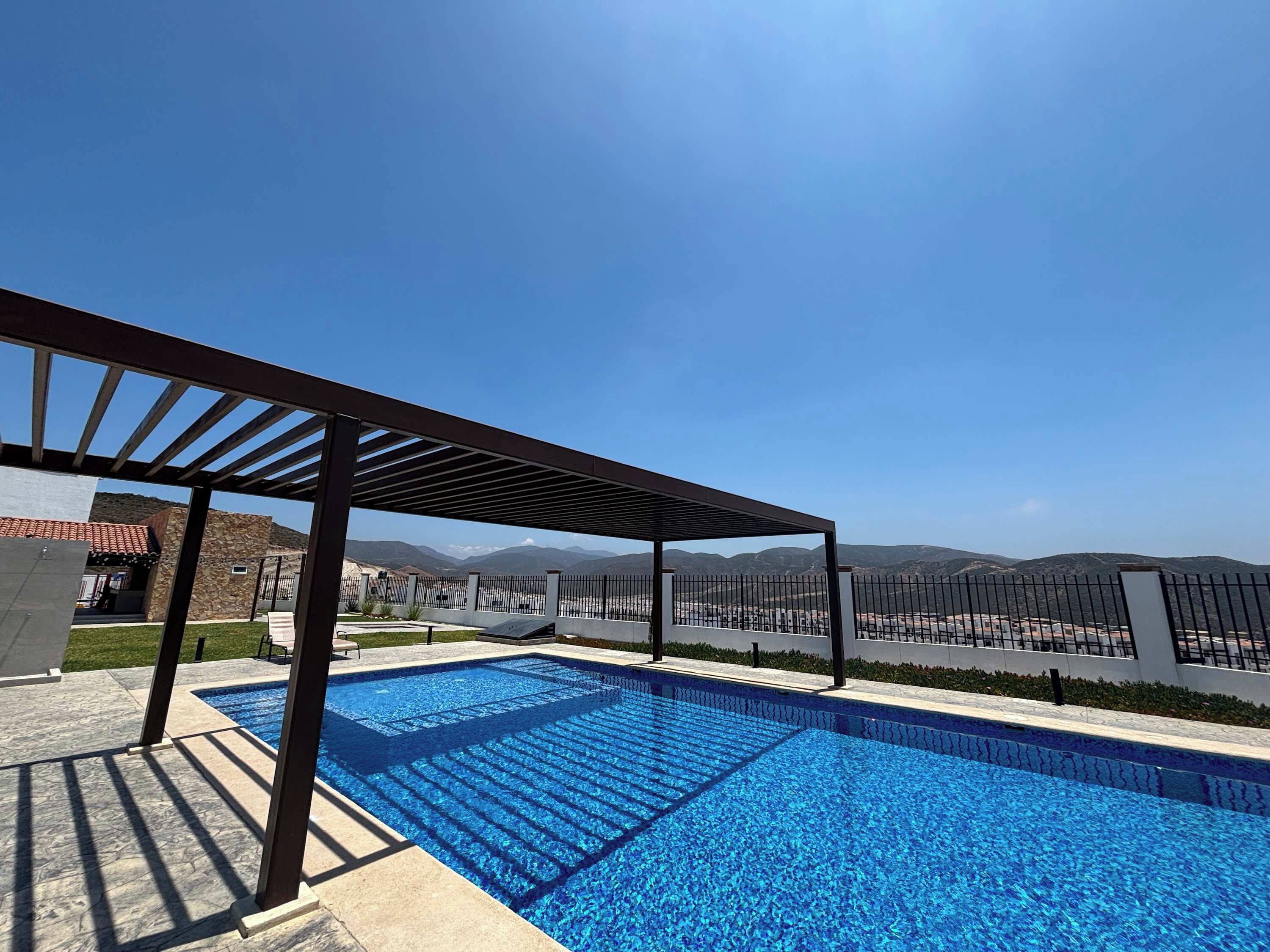A swimming pool with a pergola structure, surrounded by a fence and a view of distant mountains.