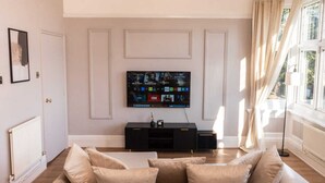 A living room with a large flat-screen TV mounted on the wall, a black entertainment unit, and a beige sofa with multiple cushions.