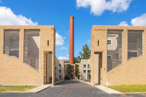 Un bâtiment doté d’une cheminée centrale, composé de deux étages en briques avec de grandes fenêtres, et une allée pavée.
