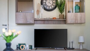 A modern living room with a flat-screen TV, wooden cabinets, a wall clock, decorative leaves, a potted plant, and a vase with tulips.