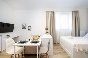 Habitación de hotel moderna y minimalista con decoración blanca, una zona de comedor con mesa y sillas, una cama con cubrecama blanco y un televisor montado en la pared.