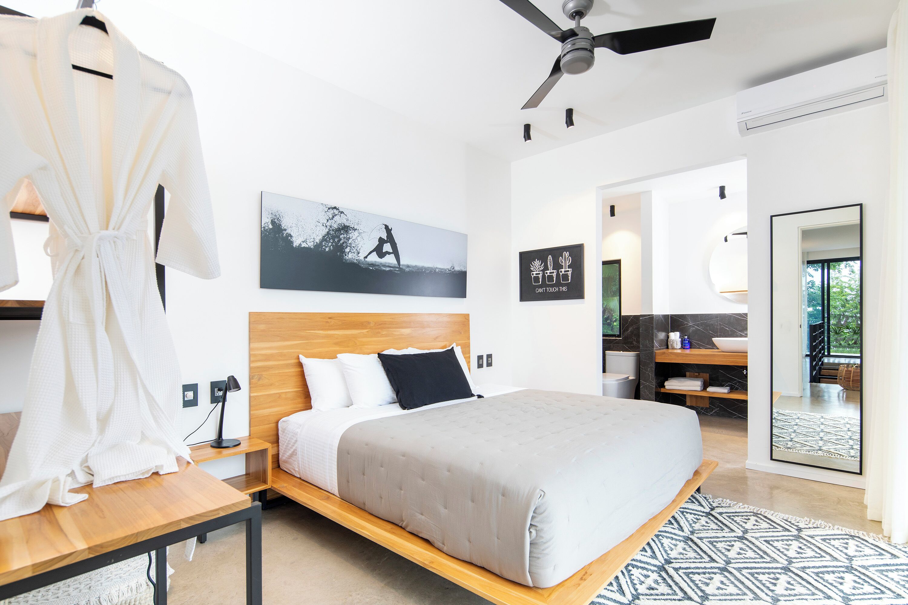 A modern hotel room with a wooden bed, a white robe, a black and white photograph on the wall, and a bathroom with a large mirror.