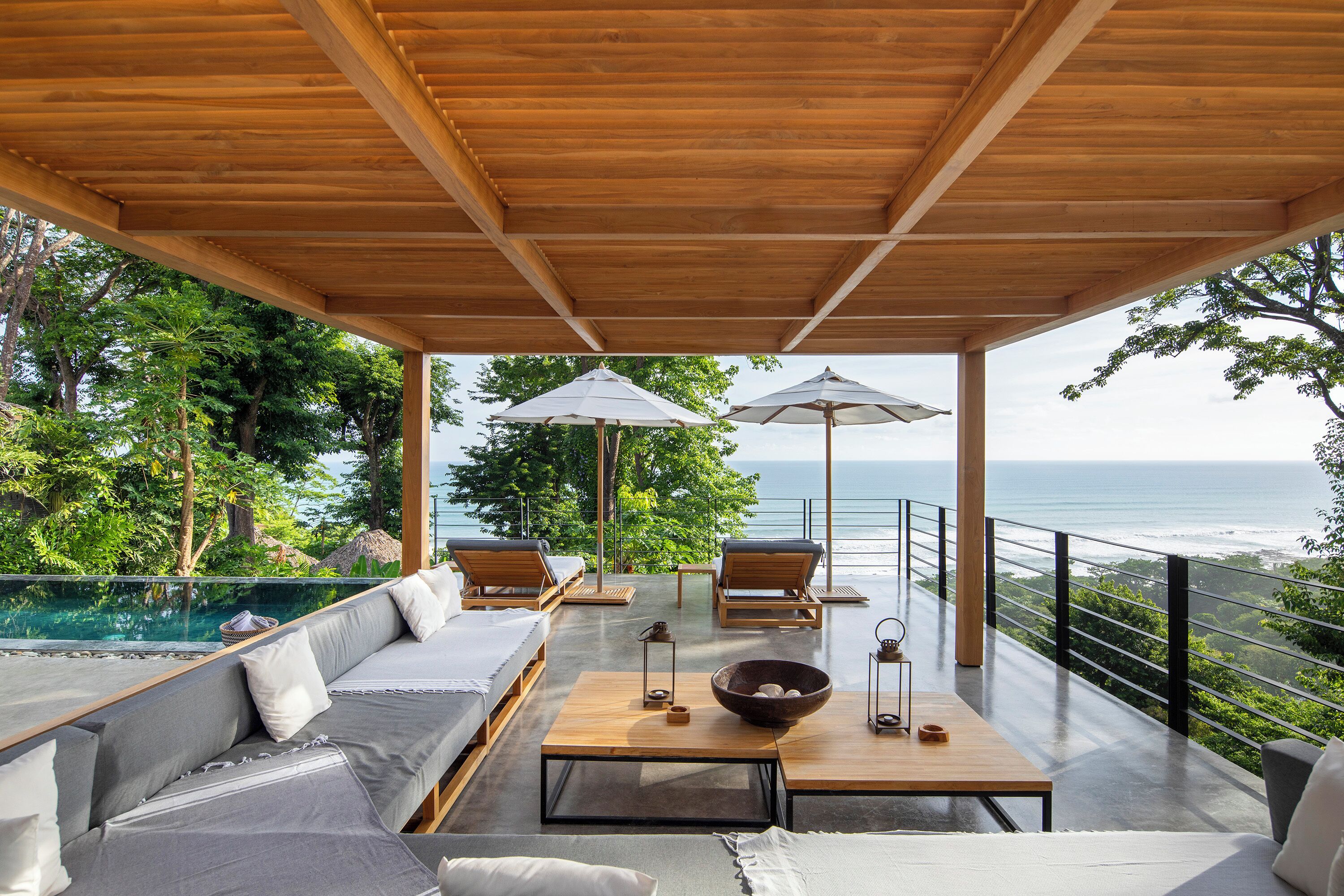 Un área de estar al aire libre techada, con piscina, mobiliario de madera y vista al océano.