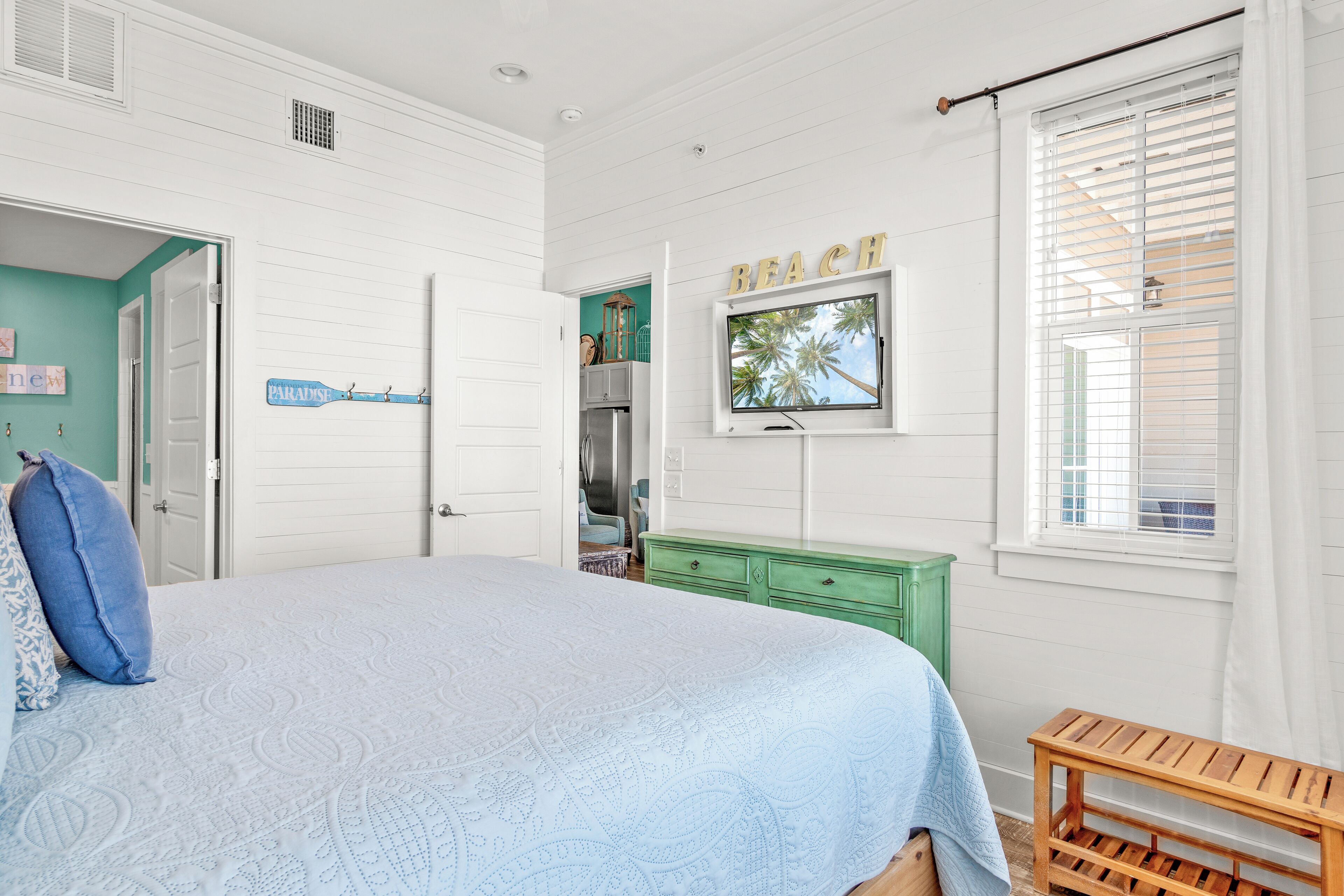 A bedroom with a bed, a green dresser, a wooden bench, and a window with blinds.