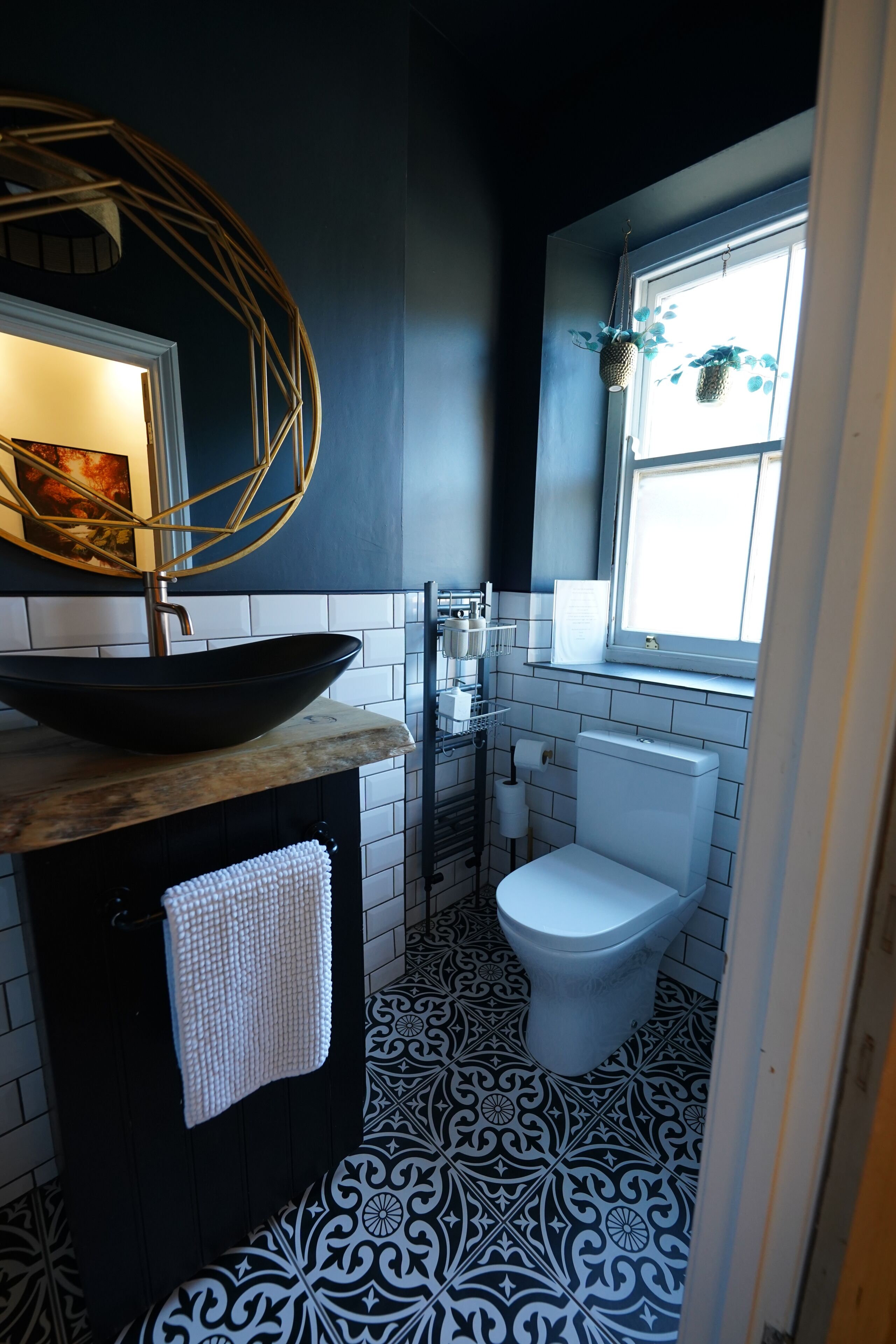 A bathroom with a black and white patterned floor, a white toilet, a black sink with a white towel, and a window with hanging plants.