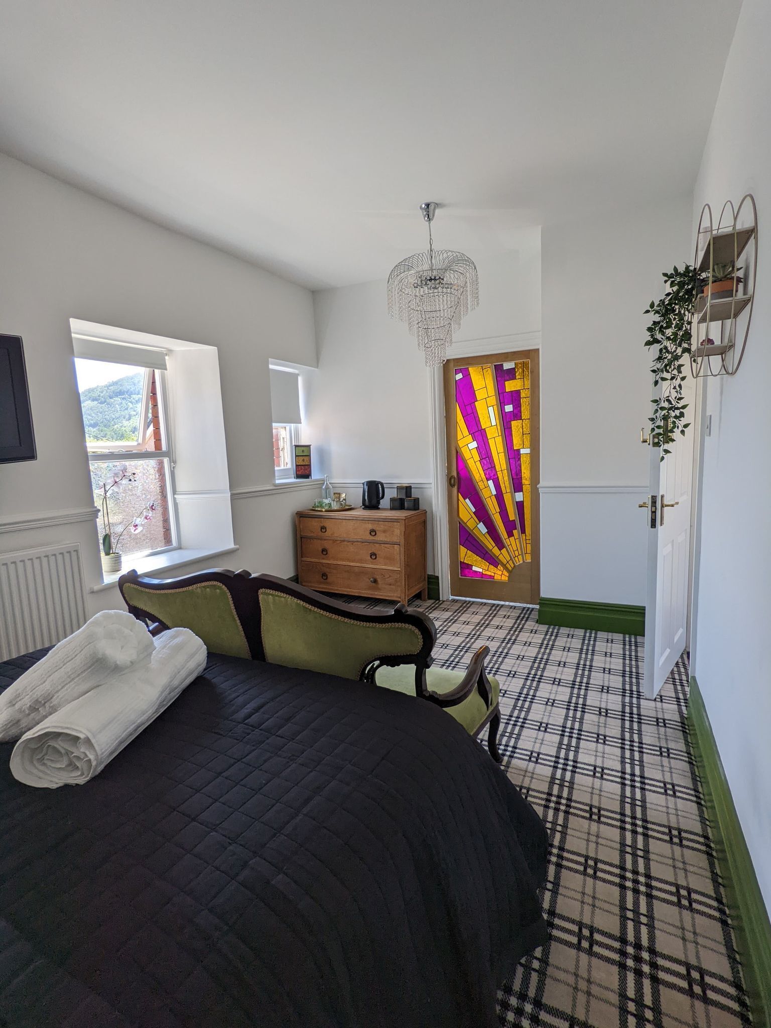 A bedroom with a bed, a dresser, a chandelier, a plaid-patterned floor, and a door with a colorful stained glass design.