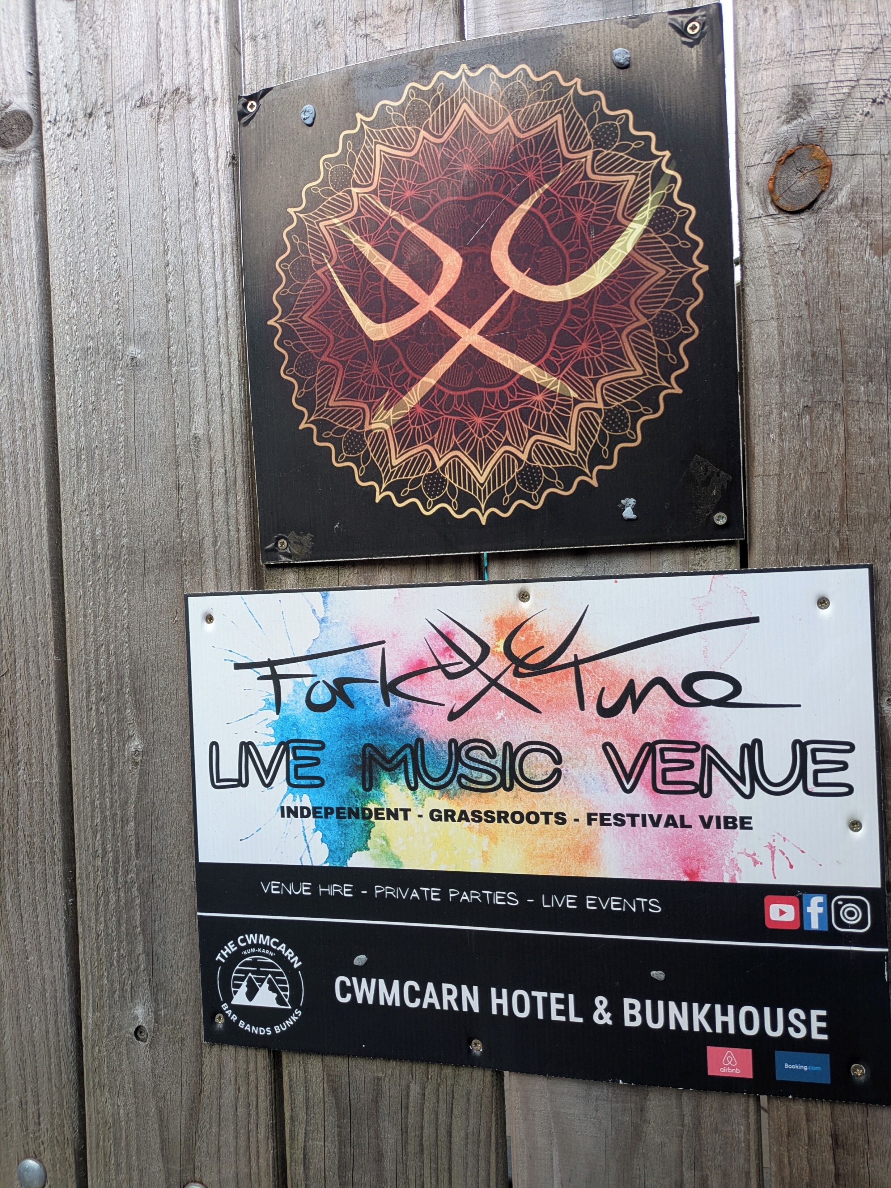 Dos carteles en una valla de madera: uno para un local de música en vivo y otro para el Cwmcarn Hotel & Bunkhouse.