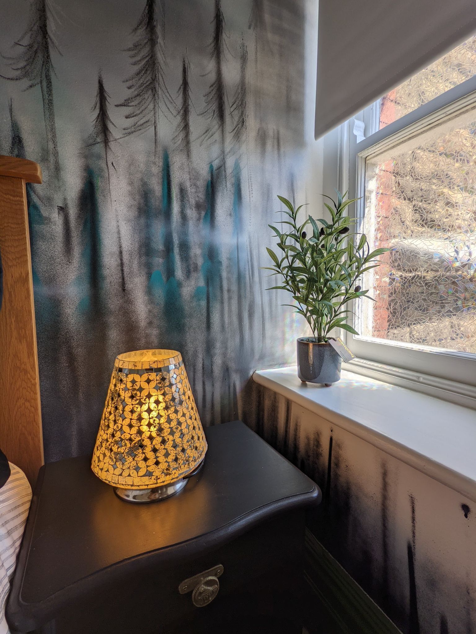 Eine Lampe auf einem schwarzen Tisch, ein Blumentopf an einem Fensterbrett und eine Wand mit einem Waldmotiv.