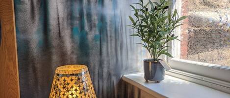 A lamp on a black table, a potted plant on a windowsill, and a wall with a forest pattern.