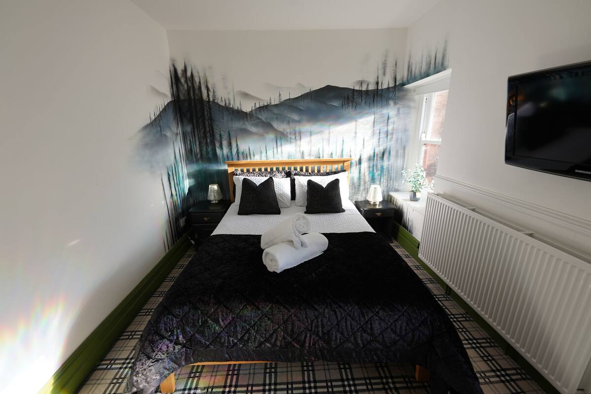 A bedroom with a bed, a TV, a window, and a wall painting.