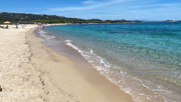 Una spiaggia con acque cristalline, litorale sabbioso e colline in lontananza.