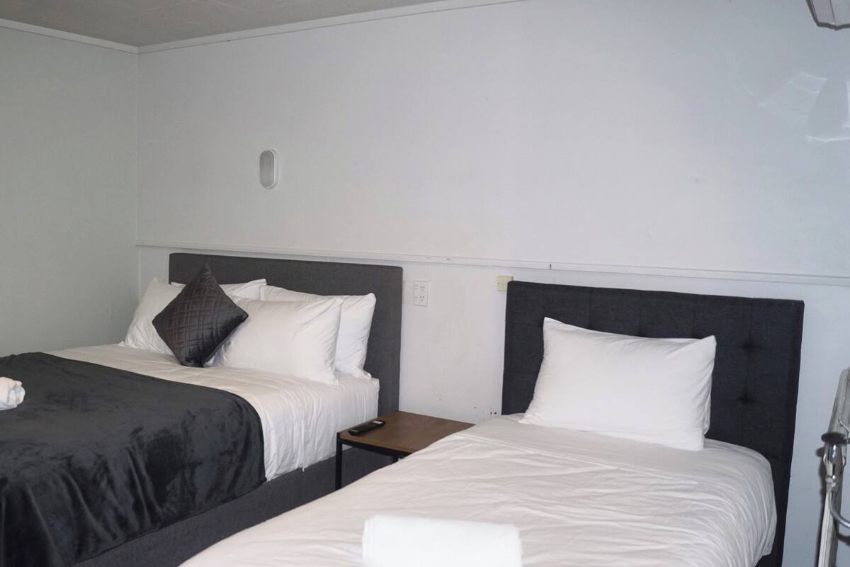 A hotel room with two beds, each with a black headboard and white bedding. There is a small wooden nightstand between the beds, and a remote control is visible on it.