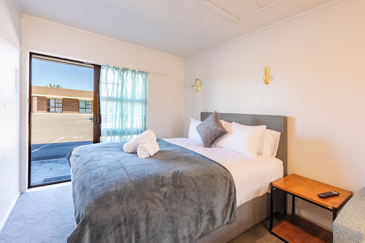 A hotel room with a bed, a small wooden nightstand, a window with curtains, and a view of the outside.