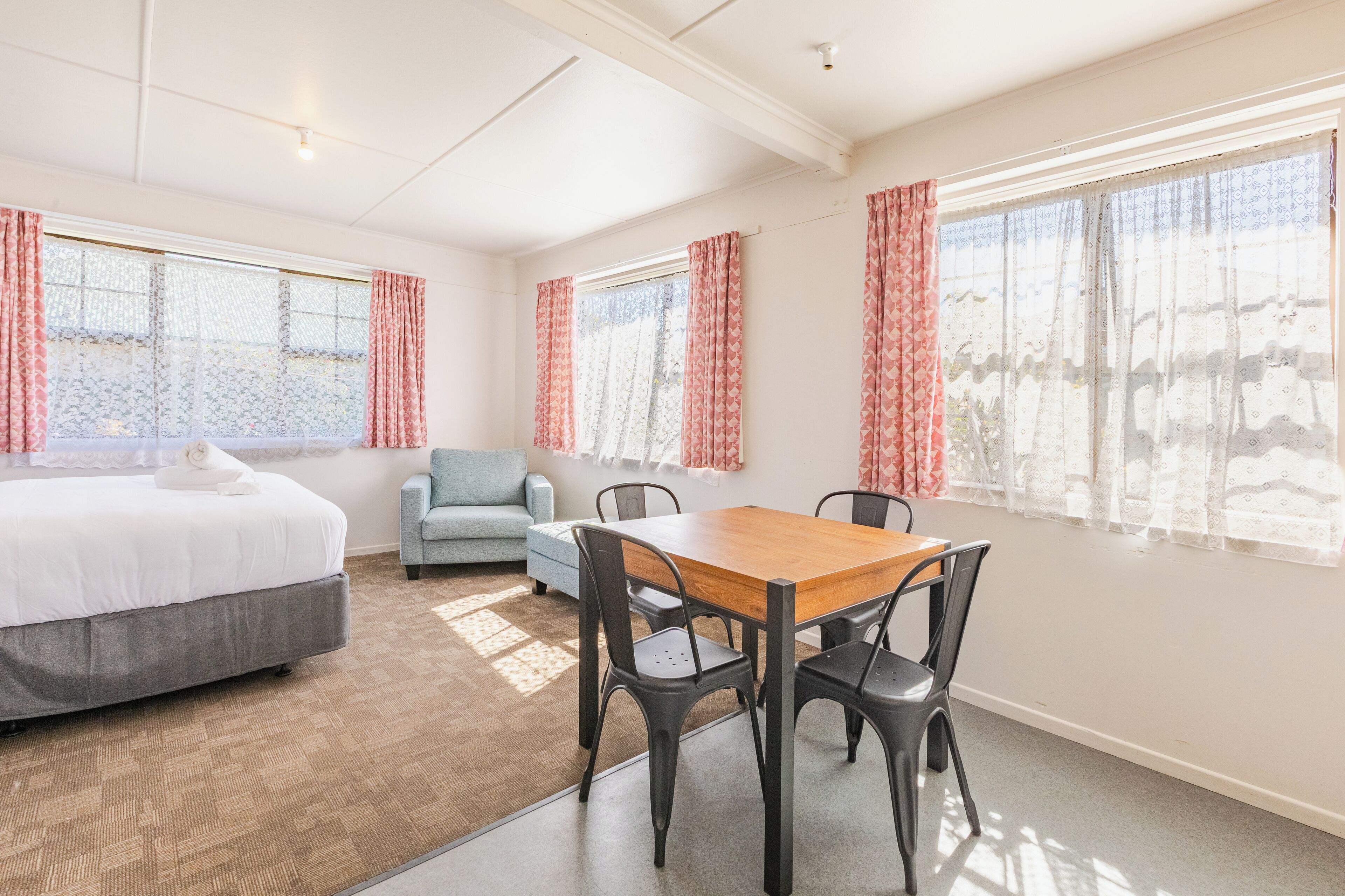 A hotel room with a bed, a small table with chairs, a sofa, and large windows with curtains.