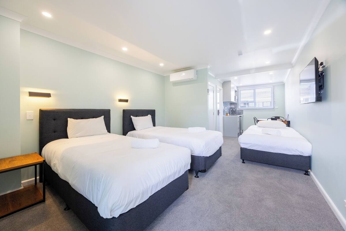 A hotel room with two beds, a small table, and a kitchenette in the background.