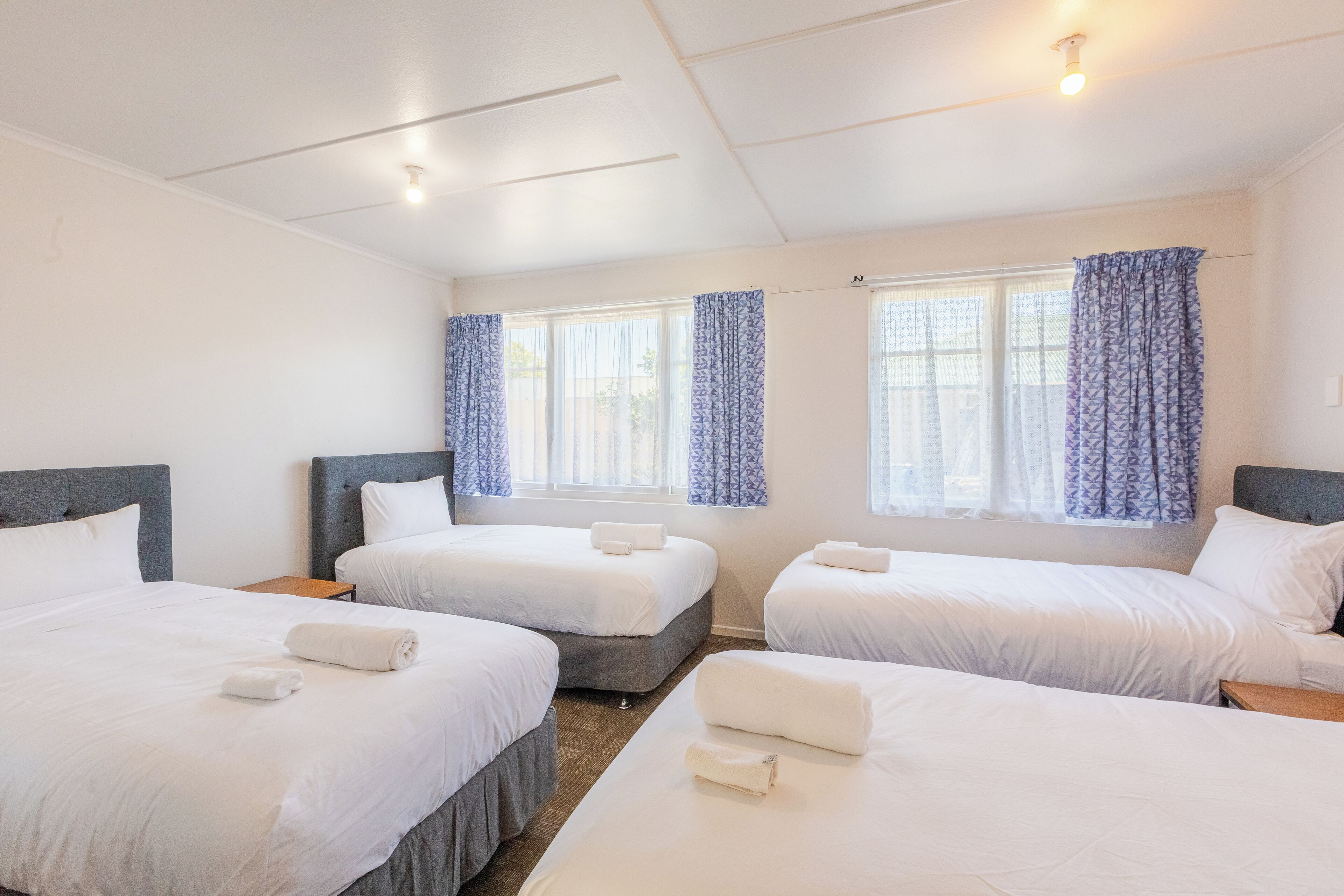 Habitación de hotel con tres camas, cada una con ropa de cama y toallas blancas, un ventilador de techo y ventanas con cortinas azules.