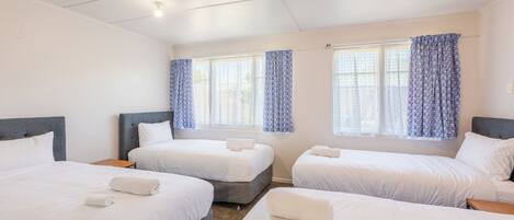Habitación de hotel con tres camas, cada una con ropa de cama y toallas blancas, un ventilador de techo y ventanas con cortinas azules.