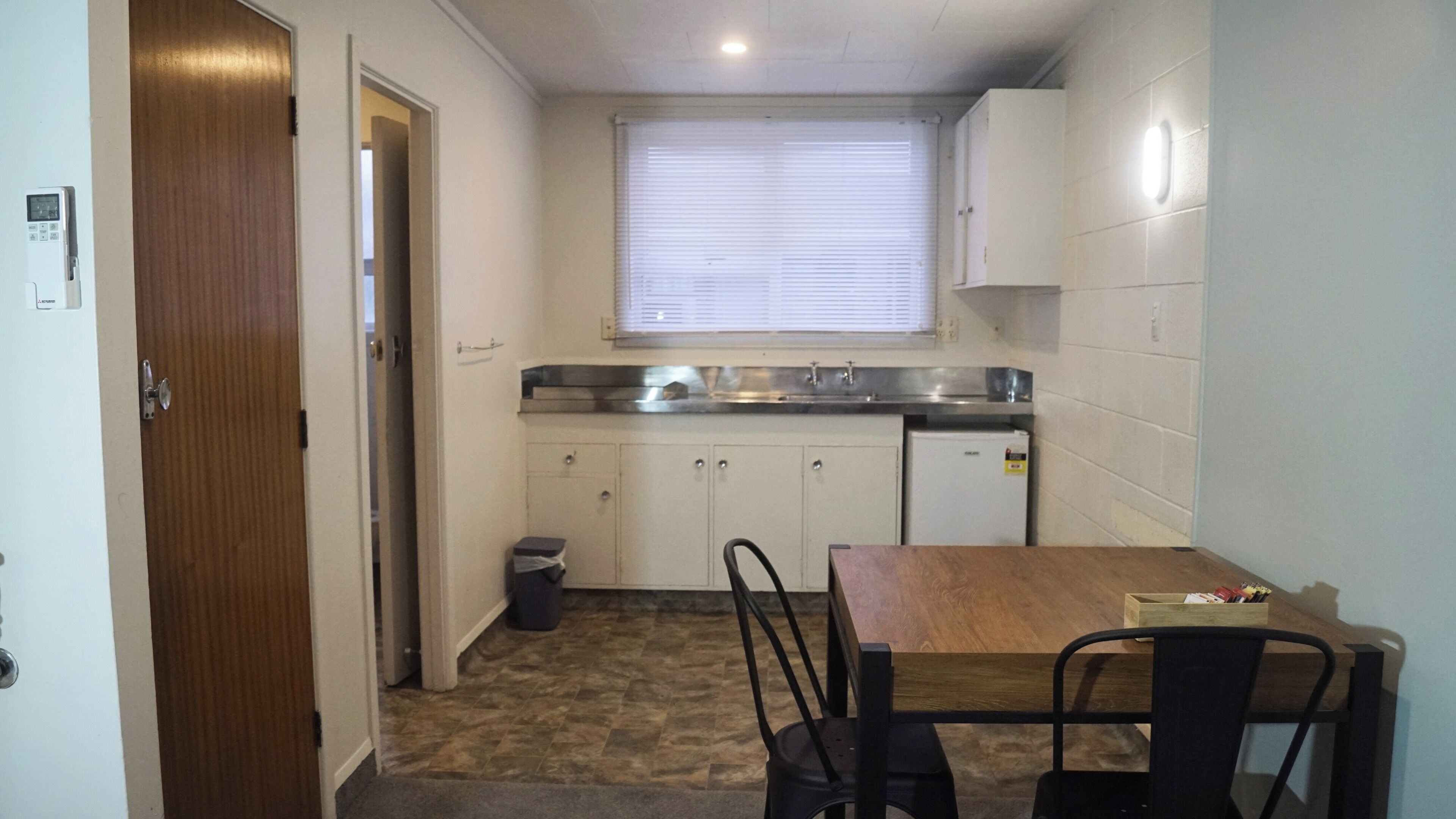Une cuisine de taille réduite, avec une table et des chaises en bois, une fenêtre avec des stores et une petite poubelle.