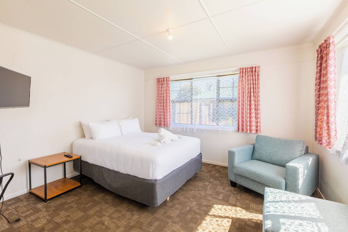 A bedroom with a bed, a small table, a chair, and a window with curtains.