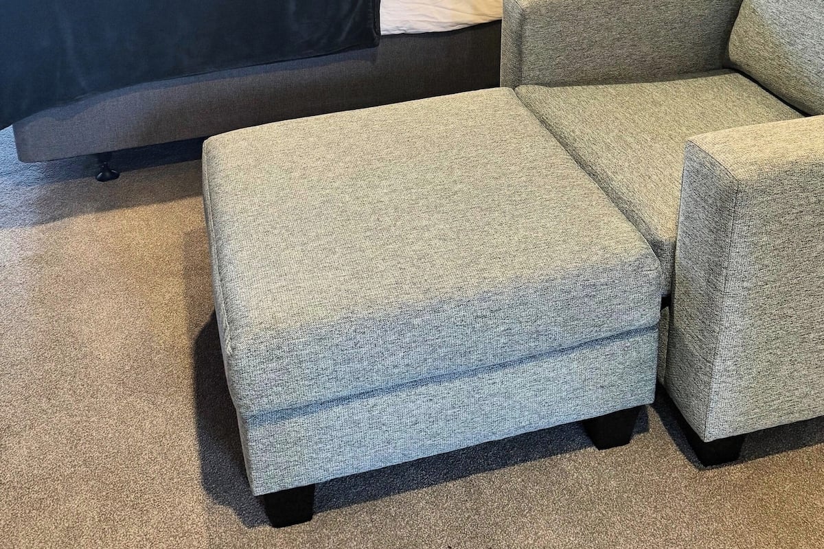 A hotel room with a bed, a grey armchair, and a matching ottoman.