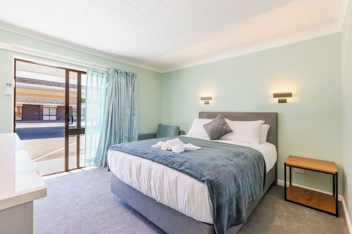A bedroom with a bed, a nightstand, a window with curtains, and a view of the outside.